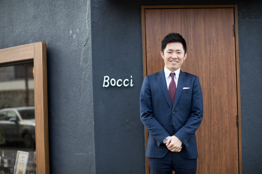 Steak&Wine Bocciの入り口ドアの横に立つ、スーツ姿の寺阪渉氏。
