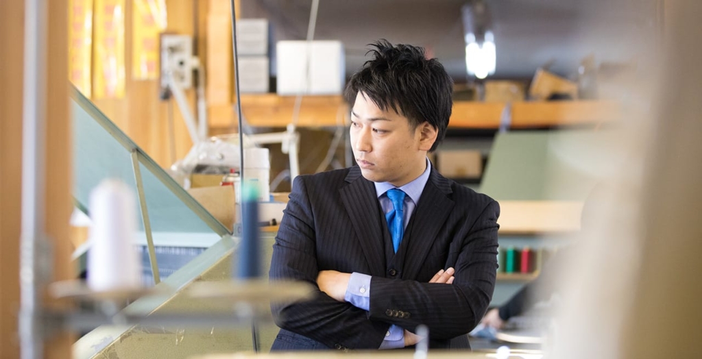 縫製工場内で腕を組むスーツ姿の笏本達宏氏。