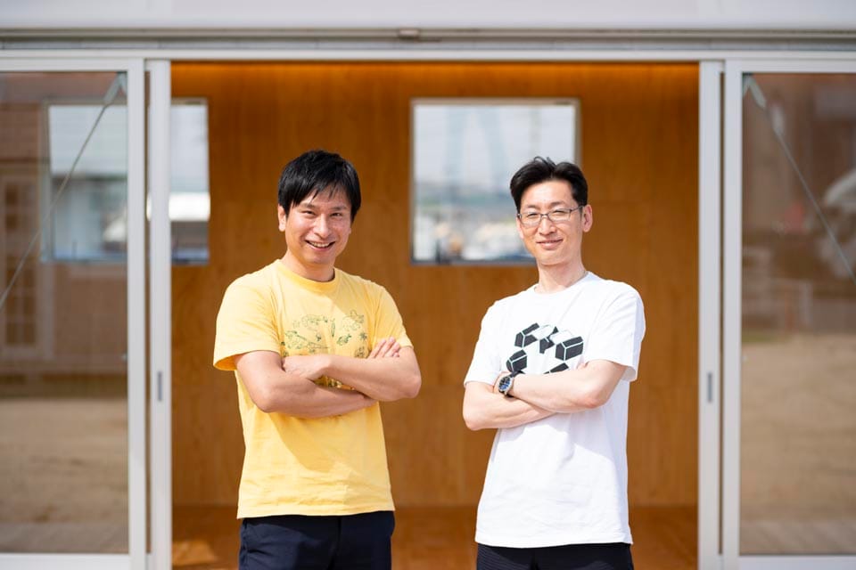 小屋の前で腕を組んで並んで立つ、植田板金店の植田博幸社長とインタビュアー。