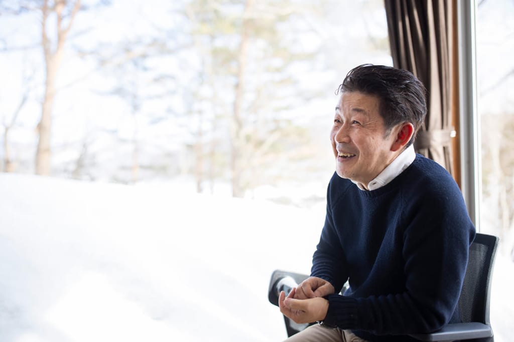 院庄林業の武本哲郎氏が、雪景色の窓を背景に椅子に座り、笑顔でインタビューに答えている。