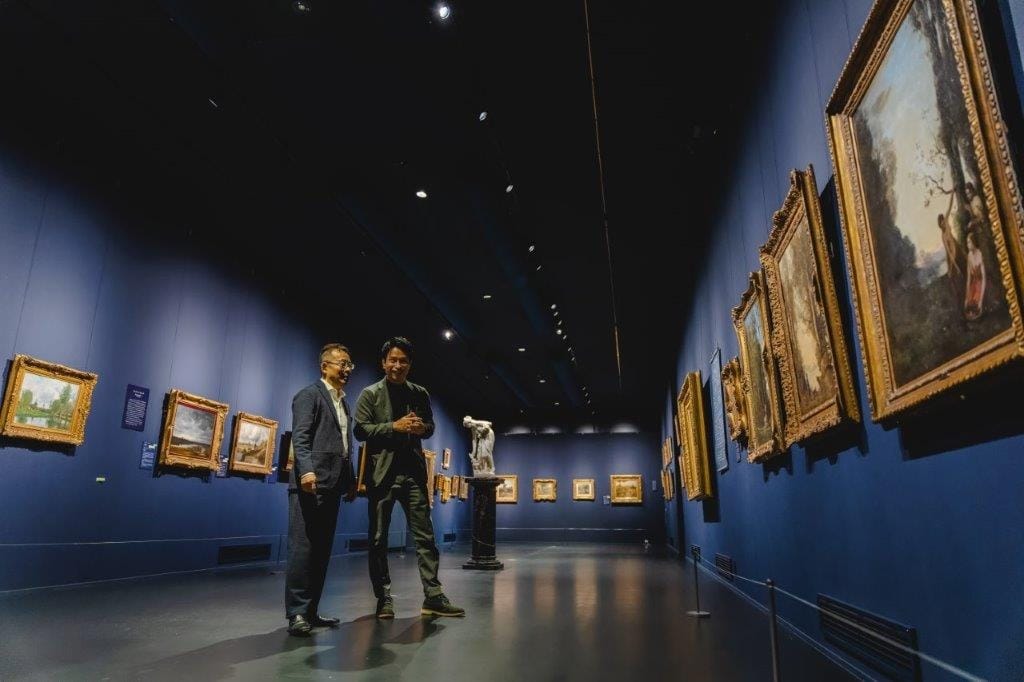 青い壁のアートギャラリーで壁にかけられた絵画を見ながら歩く山田英生社長とインタビュアー。