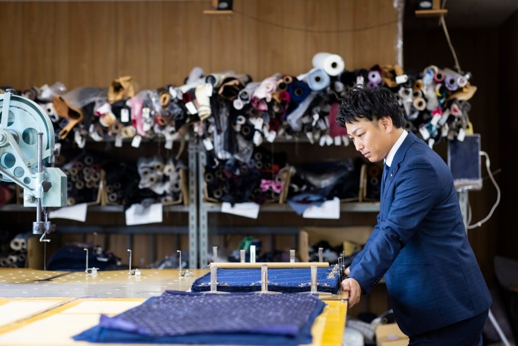 スーツ姿の笏本達宏氏が、工場内の作業台で青い柄の生地を裁断している様子。