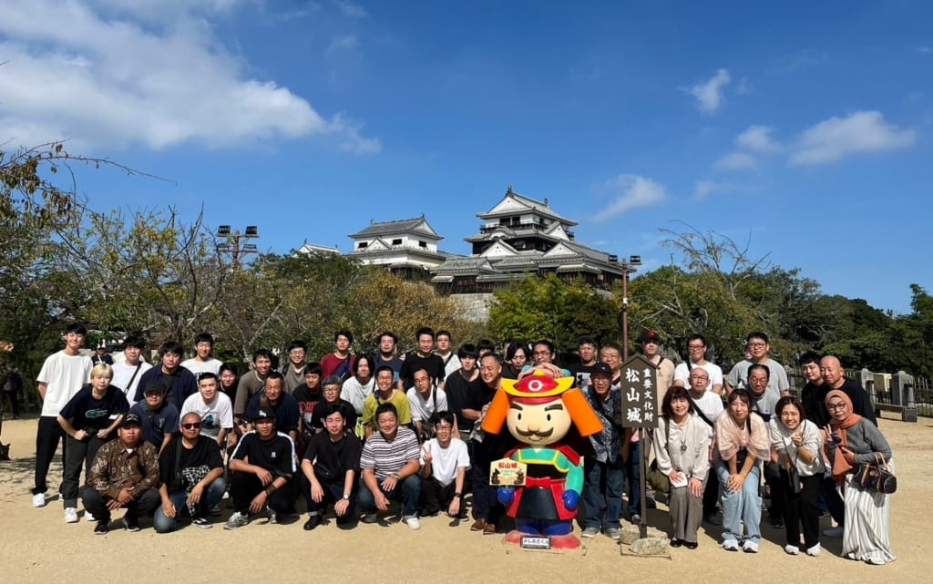 松山城をバックに、武士の格好をした二頭身のキャラクターの周りに大勢の社員が集まっている記念写真
