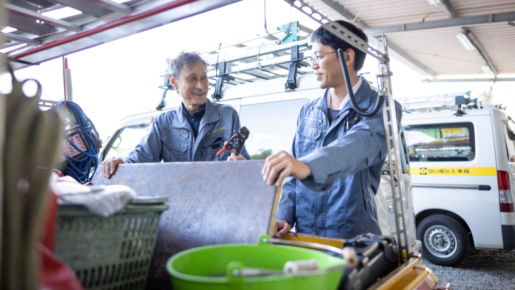 社用車の横で工具を持ち笑顔で会話する二人の社員