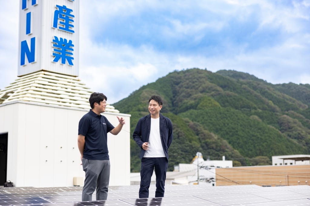 ソーラーパネルが設置された東真産業の屋上で、真庭市の山々を背景にインタビュアーと話す廣岡宣行取締役。