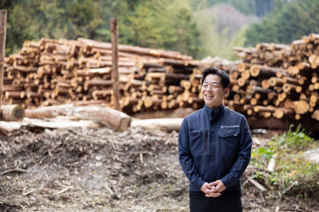 山積みの丸太を背景に、うじひら木材産業の作業着を着て立つ氏平雄人代表取締役。