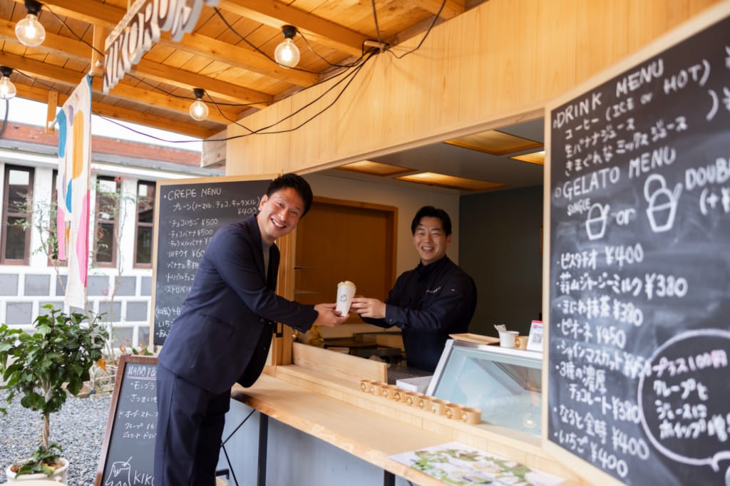 木こりのクレープ＆ジェラート店「KIKORU」のカウンターで、うじひら木材産業の氏平雄人代表取締役がインタビュアーにクレープを手渡している。