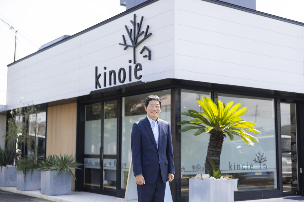 株式会社キノイエの白いモダンな社屋の前で、スーツ姿の樋口大介代表取締役が立っている。