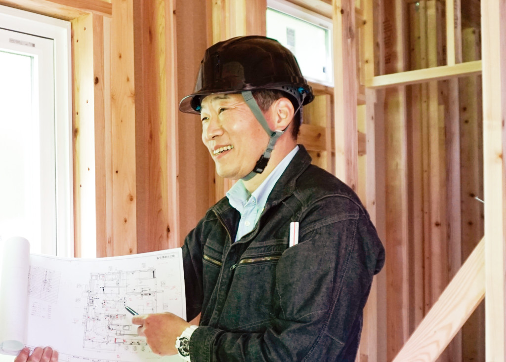 ヘルメットを着用した樋口大介代表取締役が、建設中の木造住宅の内部で、図面を指差しながら笑顔でこちらに説明を行っている。