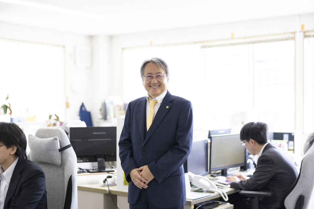 スーツ姿の男性（代表取締役 市克吉）がオフィスに立っている。背景にはデスクで作業する他の社員がいる。
