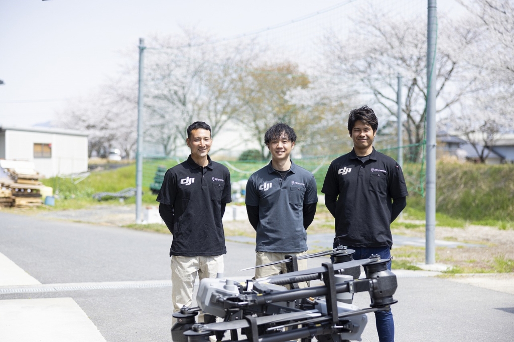 桜の見える屋外でドローンと撮影をする社員3名