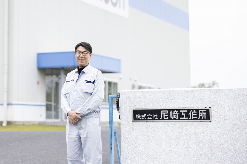 株式会社尼﨑工作所の工場前で笑顔で立つ社員、作業着姿で会社の顔となるベテラン
