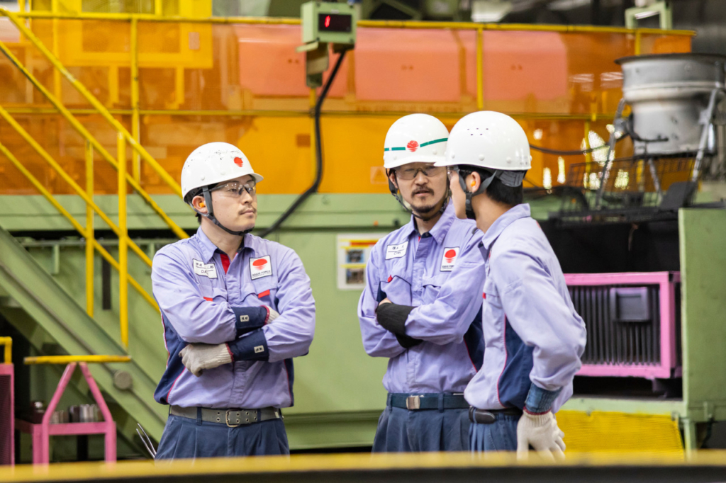 工場内の大型機械の前でヘルメットを着用し、話している3名の社員