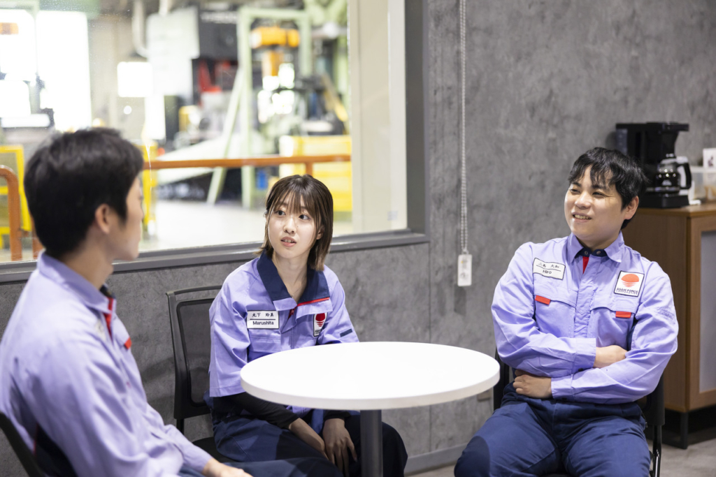 作業着を着た3人の社員が休憩室のテーブルを囲んで話している様子