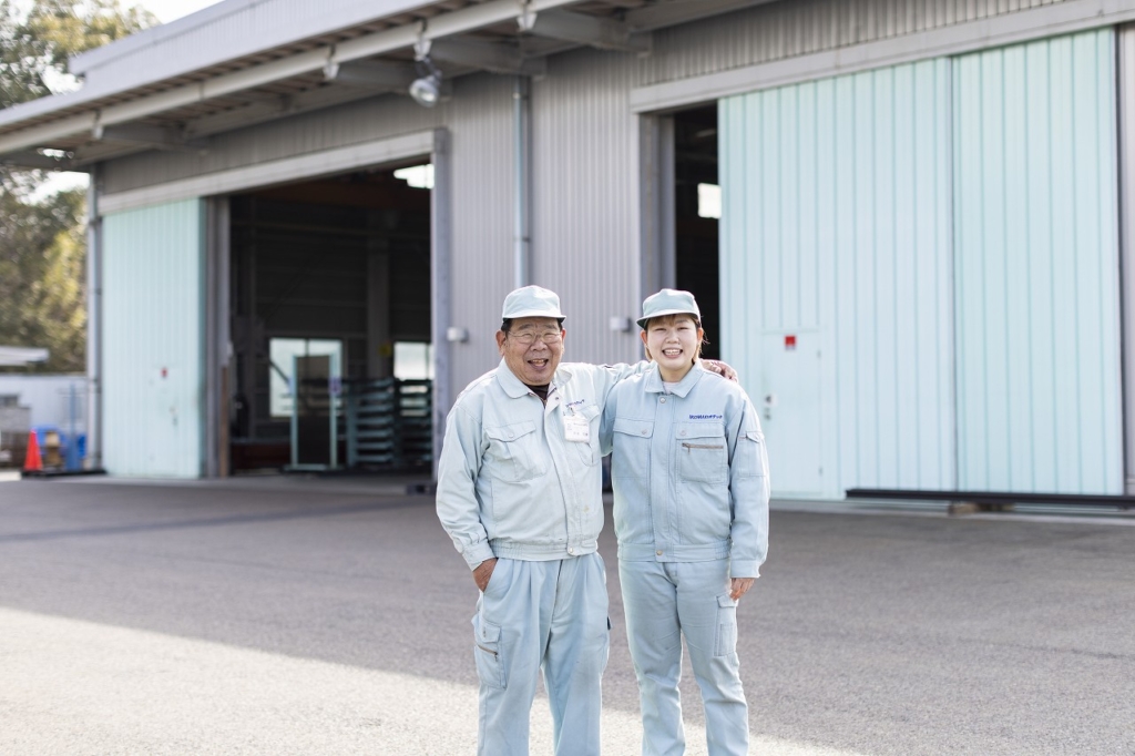 工場の外で土井さんと年の離れた男性社員が笑顔で肩を組んで並んでいる