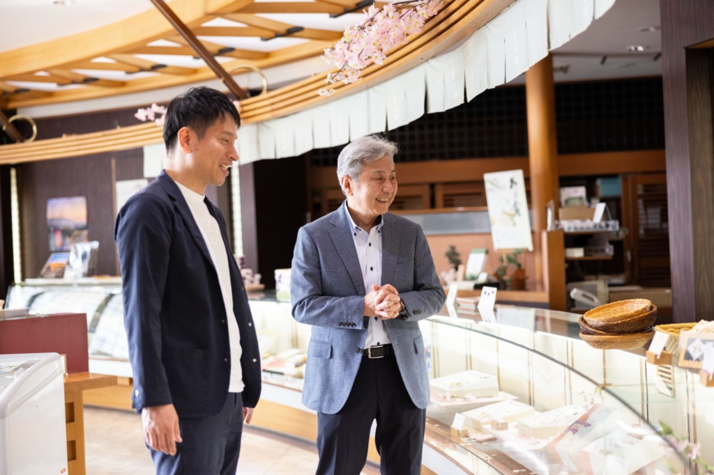 和菓子店「くらや」のショーケースの前でにこやかに話す稲葉さんと丸尾