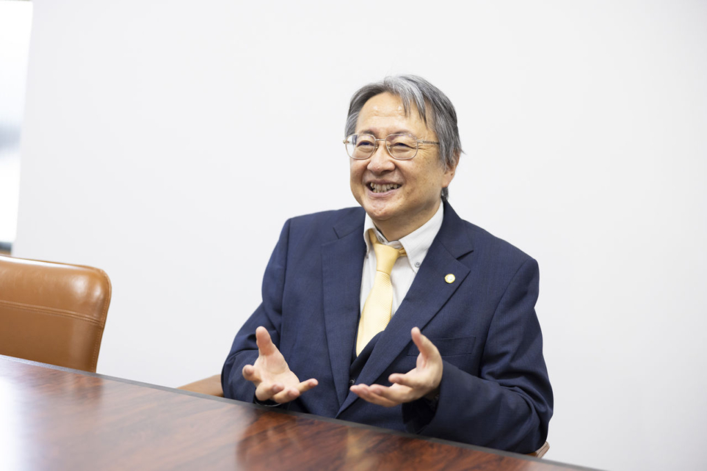 スーツ姿の男性（代表取締役 市克吉）が会議テーブルに座り、両手を広げて笑顔で話している。