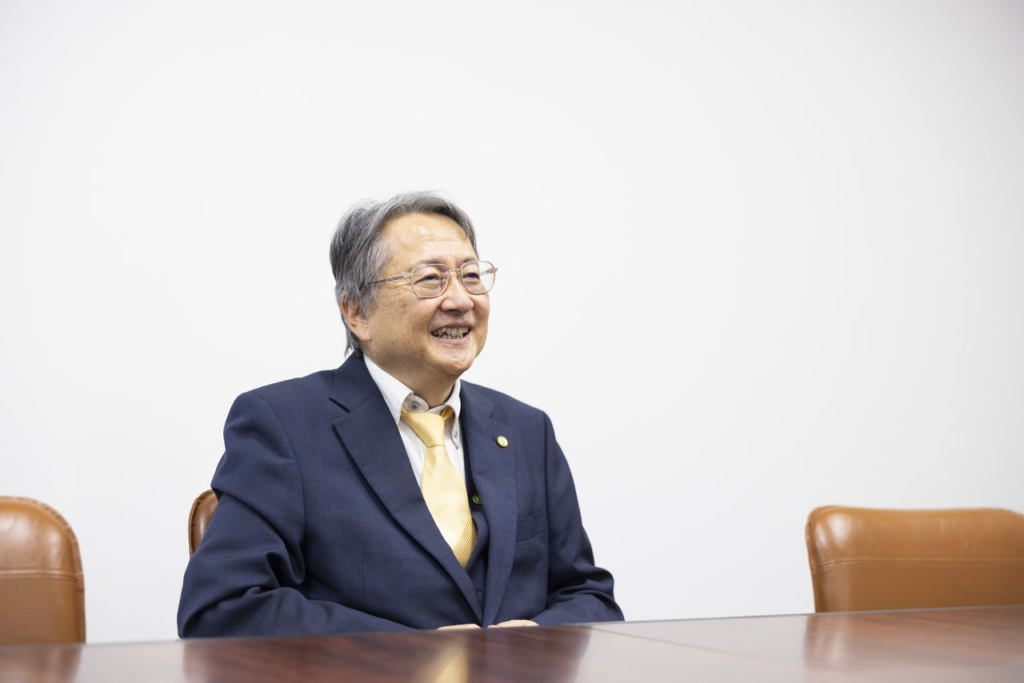 スーツ姿の男性（代表取締役 市克吉）が会議テーブルに座り、笑顔で話している。