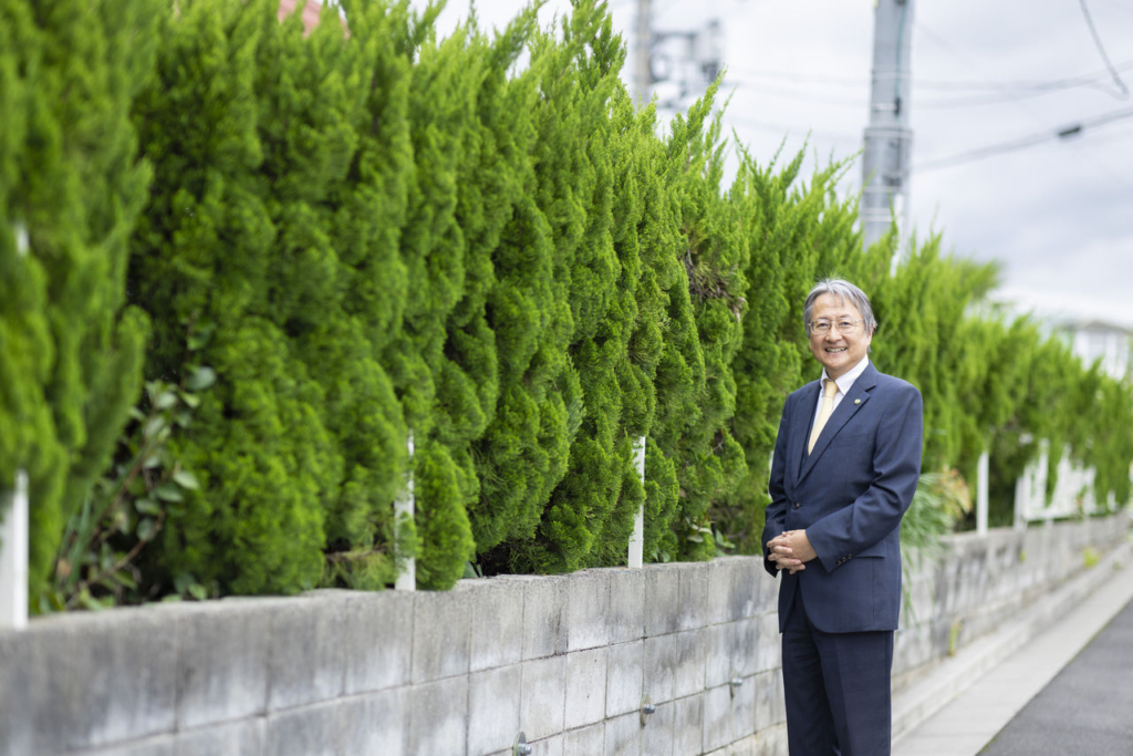 スーツ姿の男性（代表取締役 市克吉）が、緑の生垣を背景に屋外で両手を組んで立っている。