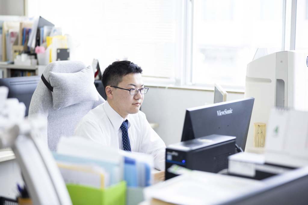 オフィスで男性社員（稲垣佑都さん）が眼鏡をかけ、デスクでPCモニターに向かって作業している。