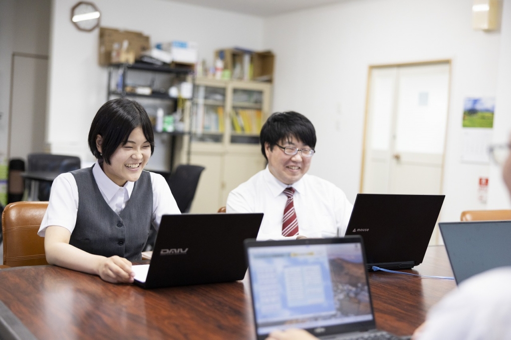 会議室で、数人の従業員が各自のノートパソコンを使いながら笑顔で話し合っている様子