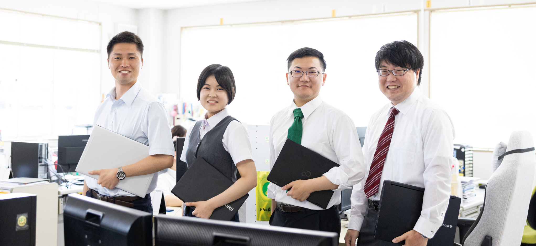 オフィス内で、ノートパソコンを抱え、笑顔で並んで立っている4人の従業員