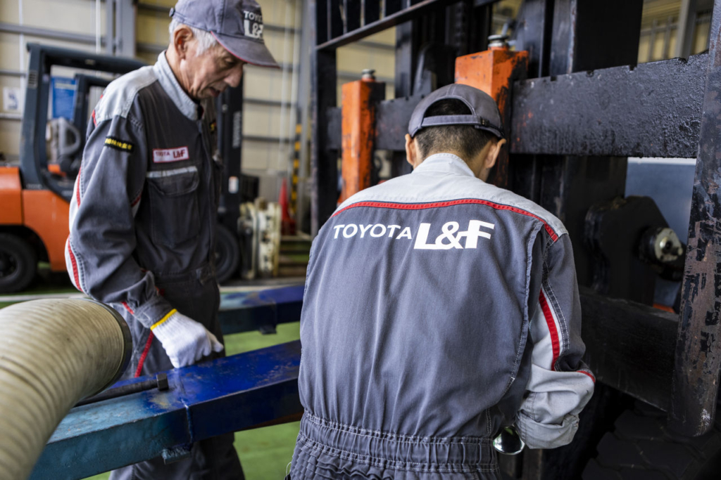 トヨタL&F岡山の整備工場で、ベテランの先輩整備士が若手スタッフにフォークリフトの整備技術を指導している様子。