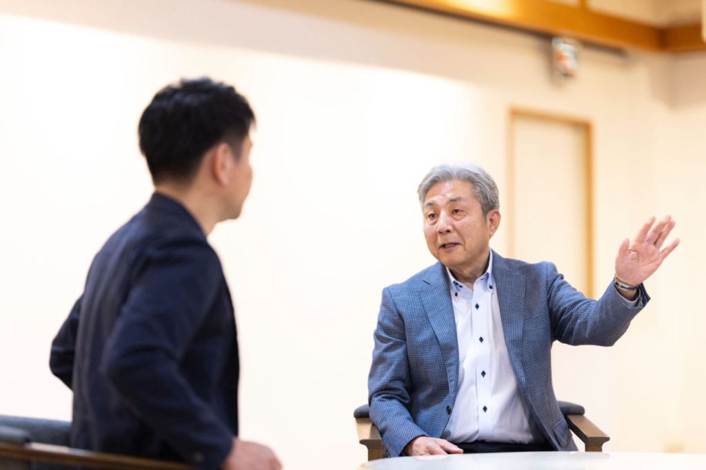 椅子に座り、右手を少し上げながら話している稲葉社長。