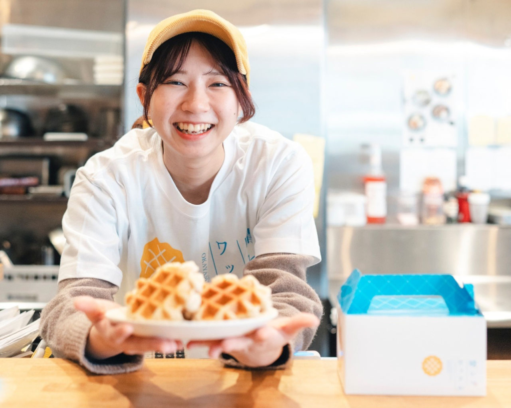ワッフル専門店の女性スタッフが、皿に乗せたプレーンワッフルを持って笑顔で立っている。
