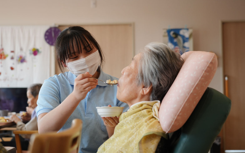 マスクをした若い女性スタッフが、椅子に座る高齢の女性利用者にスプーンで食事を運んでいる。
