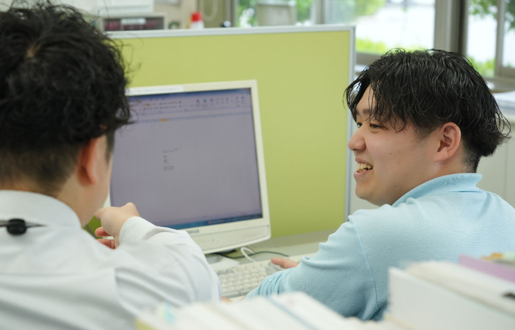 オフィスで、2人の男性スタッフがパソコンの画面を見ながら話している。