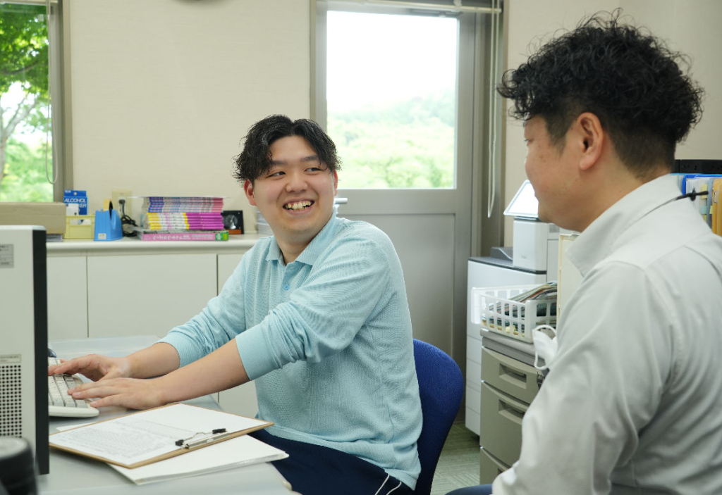 オフィスで、男性スタッフがパソコンを操作しながら、隣に座る同僚と話している。