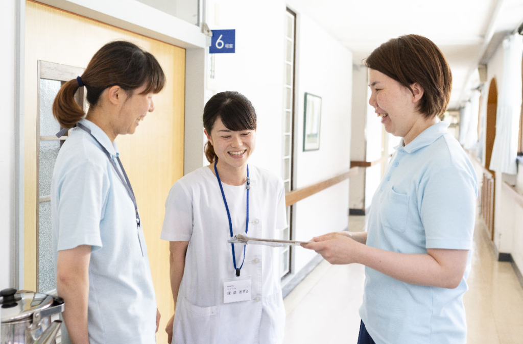 施設の廊下で、制服の違う3人の女性スタッフが書類を見ながら話している。