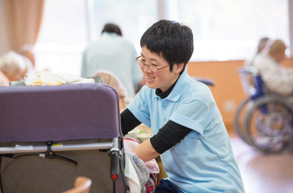 介護施設内で、女性スタッフが車椅子の準備をしている。