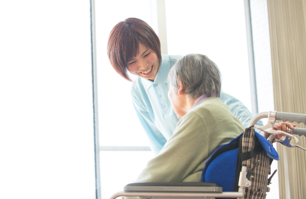 明るい窓際で、女性スタッフが車椅子に座る高齢の女性利用者に話しかけている。