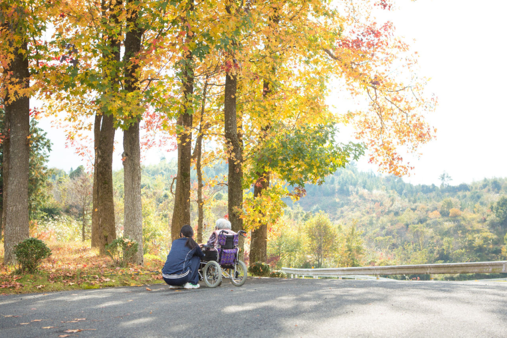 紅葉した木々が並ぶ道路で、スタッフが車椅子に座る利用者の横にしゃがんで話している。