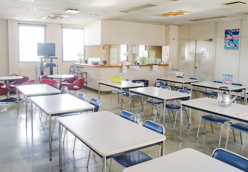 白いテーブルと青い椅子が並び、窓から光が入る社員食堂