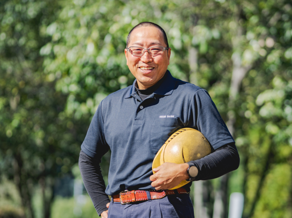 屋外の緑を背景に、黄色いヘルメットを持って立つ男性工場長