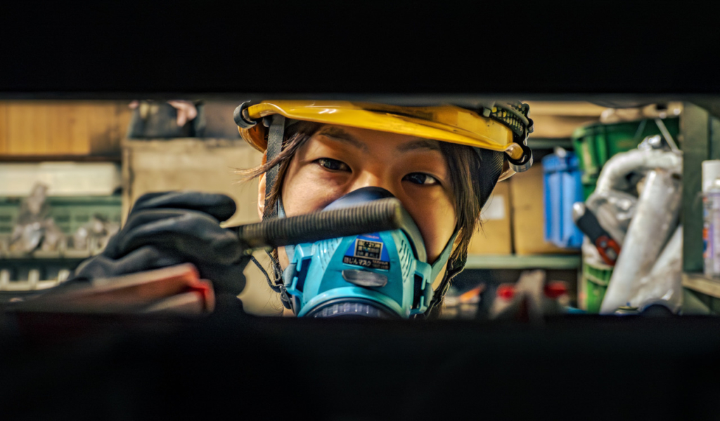 機械設備の隙間から顔を出し、防塵マスクをつけて目視点検をする作業員