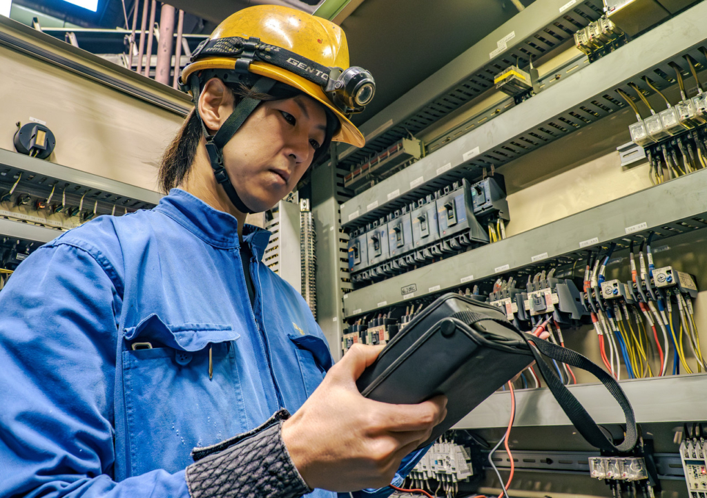 ヘルメットにライトをつけた作業員が、テスターを使って電気配電盤を点検している様子