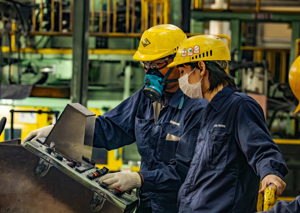 工場内の機械操作盤の前で、若手社員に指差して説明をする先輩社員