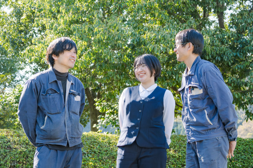 屋外の植え込みの前で談笑している作業服姿の男性2名と制服姿の女性1名