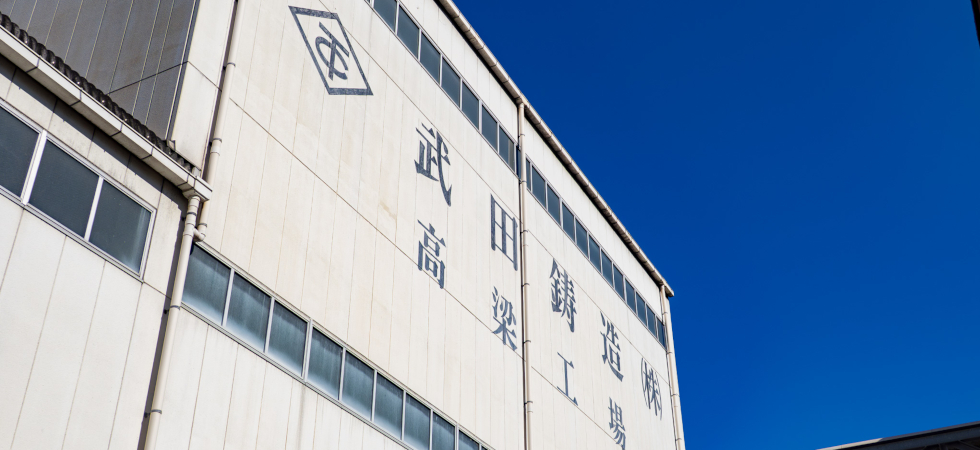 白い壁面に「武田鋳造（株）高梁工場」の文字とロゴが描かれた工場外観