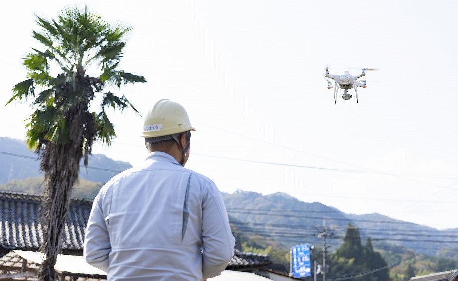 飛行中の白いドローンと、それを操縦する作業員の後ろ姿