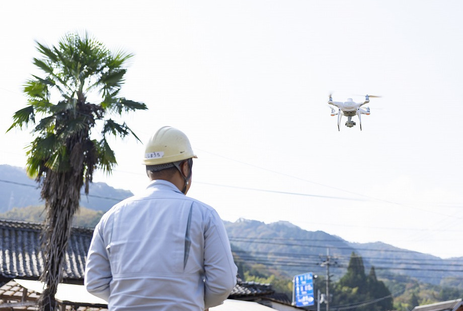 飛行中の白いドローンと、それを操縦する作業員の後ろ姿