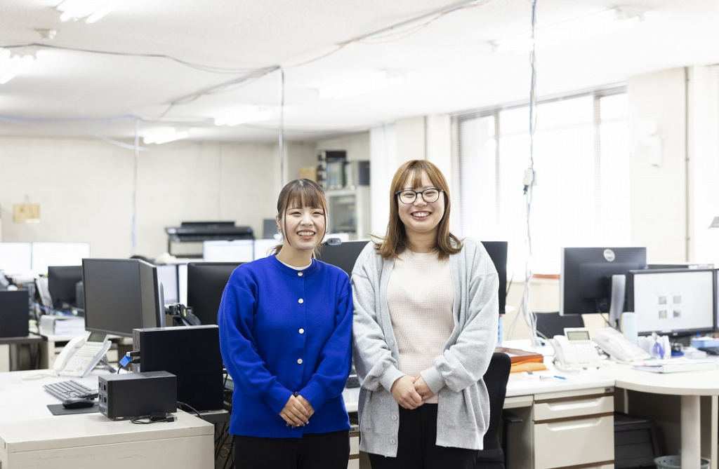 オフィス内で並んで立ち、笑顔を見せる2名の女性社員