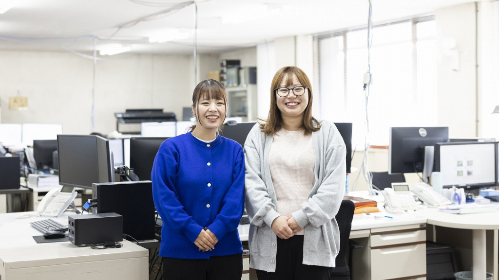 オフィス内で並んで立ち、笑顔を見せる2名の女性社員