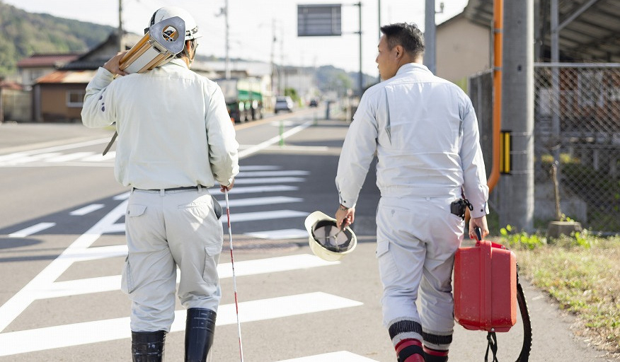 測量機材を持ち、道路を奥へ向かって歩く2名の作業員の後ろ姿