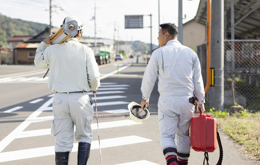 測量機材を持ち、道路を奥へ向かって歩く2名の作業員の後ろ姿