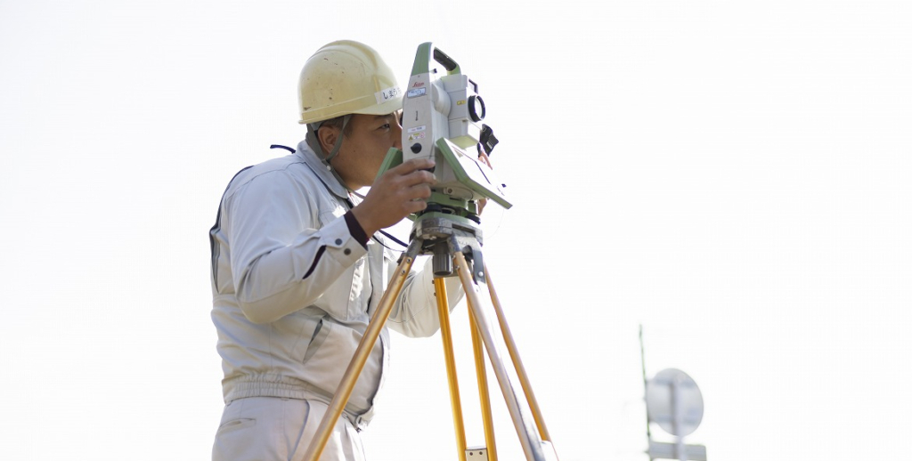 ヘルメットを着用し、三脚に据えられたトータルステーションを覗き込む男性作業員